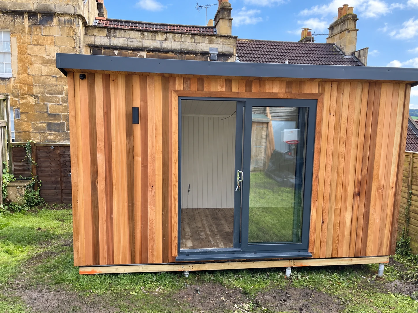 Cedar-clad Woburn eco pod with grey sliding doors, newly installed on ground screws in a walled garden